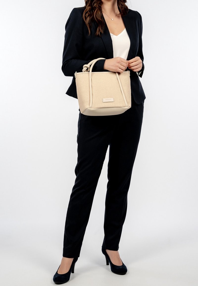 Femme en costume noir et top blanc tenant un sac à main beige texturé avec double poignée et bandoulière, debout sur fond blanc.