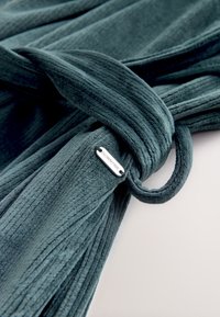 Mäkká, textúrovaná tyrkysová tkanina so zviazaným uzlom, k pásu pripevnená kovová značka s vyrytým nápisom „DOWNTIME“. Jemný povrch, rebrovaný dizajn.