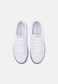 Sneakers in tela bianca con punta tonda, suola nera a contrasto e lacci bianchi. Presentano un piccolo logo in rosso e blu sulla linguetta.