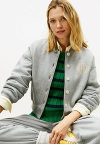 Mujer sentada con chaqueta gris abotonada sobre un suéter verde, pantalones de chándal grises, puños de camisa con rayas amarillas y zapatillas amarillas.