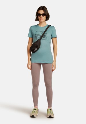 Vrouw in een teal graphic t-shirt, mauve legging, zonnebril, zwarte schoudertas en beige sneakers met neon groene veters, op een witte achtergrond.
