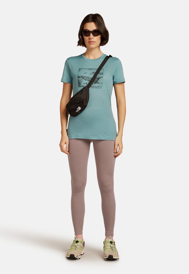 Mujer vistiendo una camiseta gráfica en color azul verdoso, leggings malva, gafas de sol, bolso cruzado negro y zapatillas beige con cordones de neón verde sobre un fondo blanco.