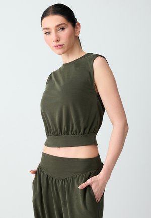 Kobieta o ciemnych włosach związanych z tyłu, ubrana w oliwkowozielony krótki top bez rękawów i dopasowane spodnie z wysokim stanem, z rękami w kieszeniach.