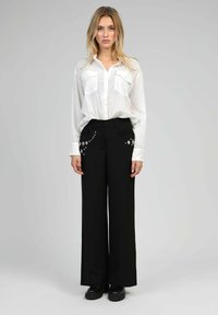 Chemise blanche à manches longues avec deux poches poitrine, associée à un pantalon ample noir avec des broderies florales au niveau des hanches. Chaussures noires.