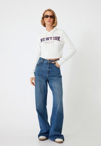 Witte geribde hoodie met de tekst "NEWYRK", gecombineerd met wijde blauwe spijkerbroek. Casual sneakers maken de outfit compleet.