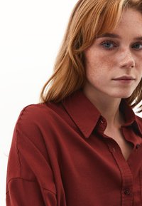 Camicia rossa con bottoni, dal colletto classico e maniche lunghe. Tessuto morbido mostrato in primo piano, con una finitura testurizzata e sottili dettagli di cucitura.