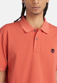 Korallfärgad polo t-shirt i bomull, med en knäppt krage, korta ärmar och en broderad trädlogotyp på vänster bröst.