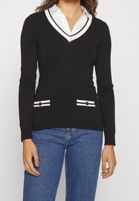 Pull en maille noir à col en V avec des bordures blanches, doté de manches longues et de deux poches avant avec des accents rayés. Porté par-dessus une chemise à col.