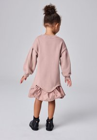 Lapsi, jolla on kiharat hiukset nutturalla, pukeutuneena pitkähihainen pölyinen vaaleanpunainen mekko, jossa on röyhelöinen helma, ja mustat nilkkurit, takaviistossa.