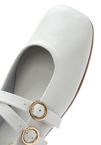 Scarpa in pelle bianca con punta arrotondata, dotata di due cinturini incrociati con accessori circolari in tono oro e suola piatta.