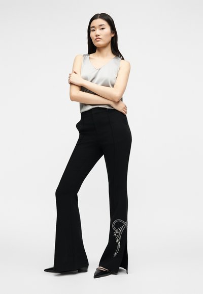 Γυναίκα στέκεται με σταυρωμένα χέρια, φοράει ασημί αμάνικη μπλούζα και μαύρο καμπάνα παντελόνι με λευκό φιόγκο και λουλουδάτη κεντητική κοντά στο τελείωμα.