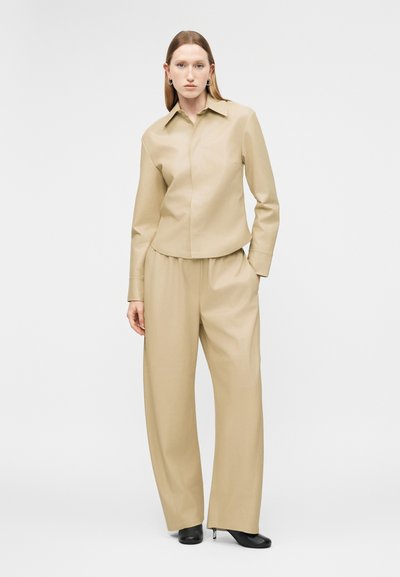 Žena stojaca, oblečená v béžovej blúzke s dlhým rukávom a golierom a v rovnakých širokých nohaviciach, na nohách čierne lodičky, na jednoduchom bielom pozadí.