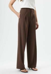Pantalon marron taille haute à jambes larges avec plis, poches latérales et tissu lisse. Associé à des sandales noires et un haut blanc.