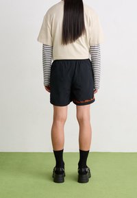 Person som står på grön golv och vänder sig mot vit vägg, klädd i en beige kortärmad över en grå-vit randig långärmad tröja, svarta shorts, svarta strumpor och skor.