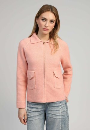 Femme aux longs cheveux blonds portant une veste zippée rose clair avec deux poches avant, associée à un jean bleu délavé.