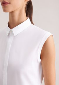 Femme portant une chemise blanche sans manches avec un col et une patte de boutonnage, visible du menton au haut du bras sur un fond neutre.