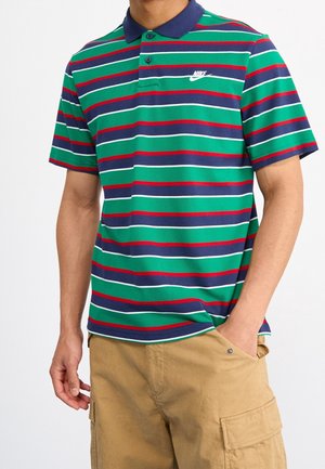 Homme portant un polo Nike rayé vert, bleu marine, rouge et blanc, associé à un short cargo beige, debout avec une main dans la poche.