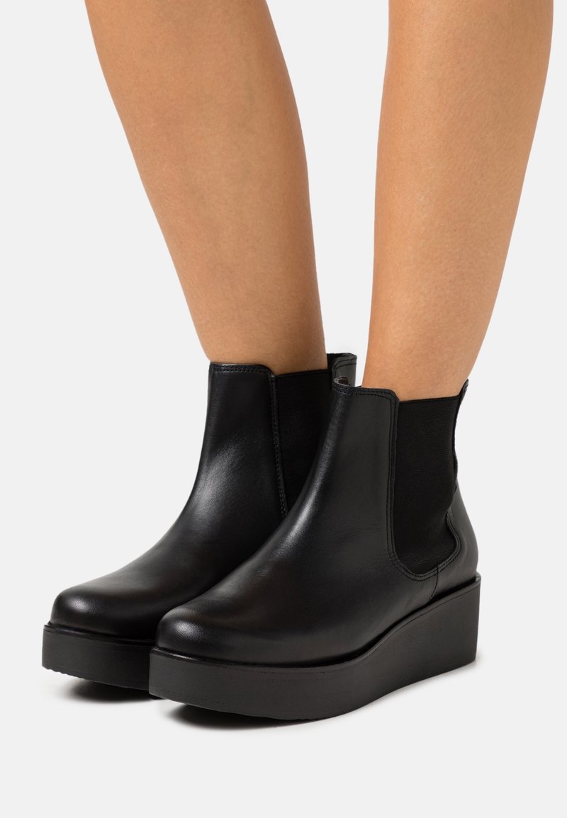 Bottes basses en cuir noir avec semelles compensées portées sur jambes nues, dotées de panneaux latéraux élastiques et d'une finition lisse et polie.