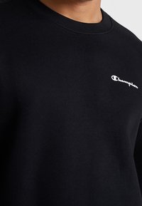 Sudadera negra de algodón, con cuello redondo y un pequeño logo blanco de Champion en el lado izquierdo delantera. Textura suave.