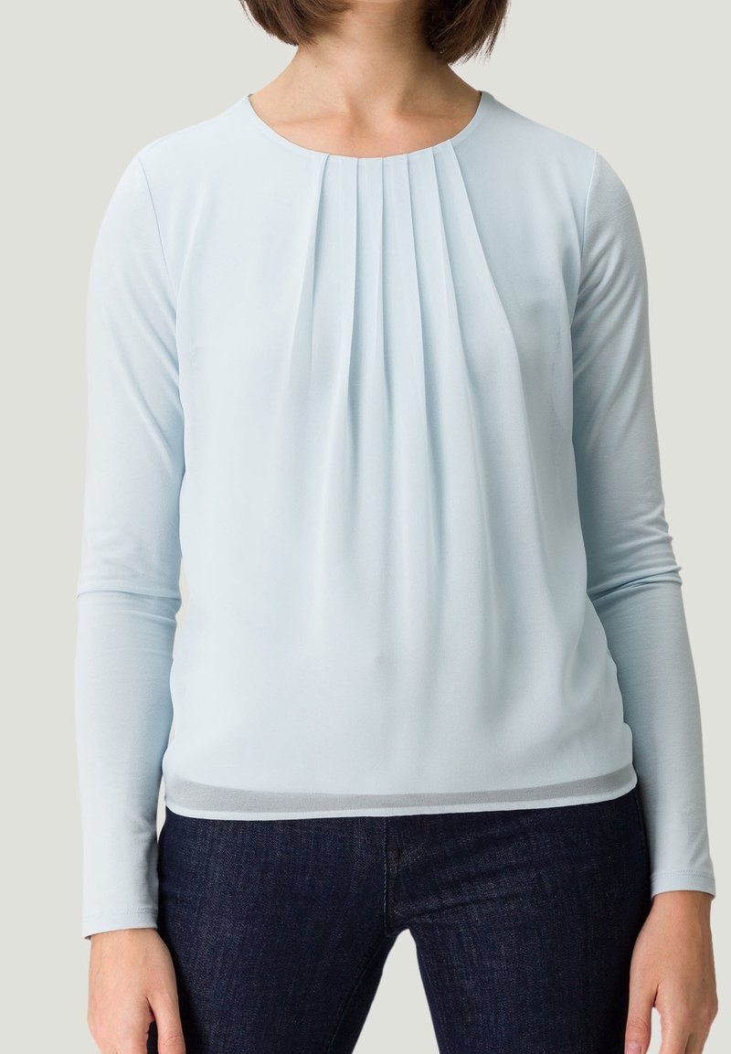 Blouse longue à manches bleues claires avec des détails plissés à l'avant, tissu lisse, encolure ronde et silhouette ajustée.