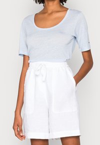 Valkoinen pellavapaita, jossa on lyhyet hihat ja joka on valmistettu pehmeästä kankaasta, yhdistettynä valkoisiin pellavashortseihin, joissa on joustava vyötärö ja huomaamattomat sivutaskut.