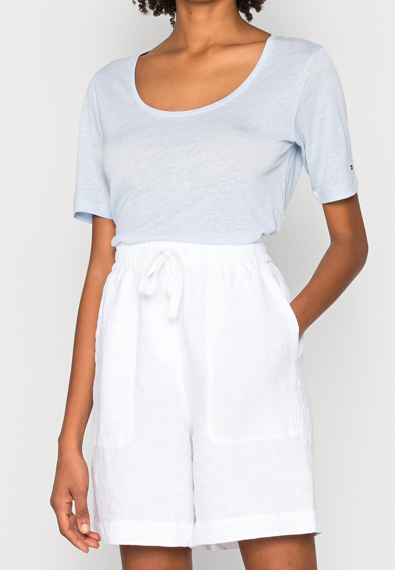 Valkoinen pellavapaita, jossa on lyhyet hihat ja joka on valmistettu pehmeästä kankaasta, yhdistettynä valkoisiin pellavashortseihin, joissa on joustava vyötärö ja huomaamattomat sivutaskut.