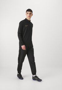 Nike zwarte lange mouw polo met een gouden logo, gecombineerd met zwarte joggingbroek. Heeft een losse pasvorm en elastische manchetten. Draag het met zwarte voetbalschoenen.