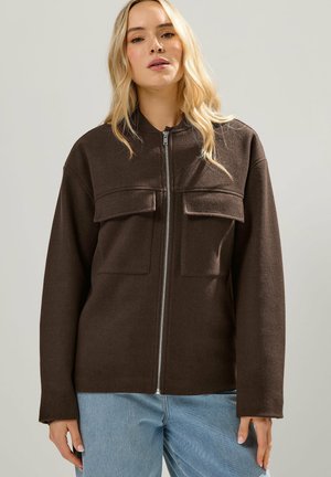 Blonde vrouw met een donkerbruine jas met rits en twee borstzakken, gecombineerd met een lichtblauwe spijkerbroek, tegen een effen achtergrond.