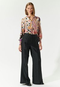 Blusa colorata e a fantasia con bottoni sulla parte anteriore e maniche lunghe; abbinata a pantaloni neri a gamba larga con vita alta e scarpe nere.