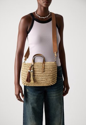 Femme portant un débardeur blanc et un jean bleu, avec un sac à main en paille tissée à poignées en cuir et décorations à pampilles porté sur l'épaule.