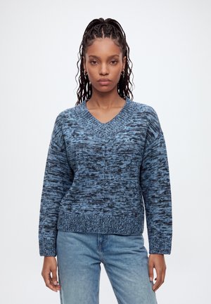 Femme aux cheveux tressés portant un pull en maille bleu à motifs et un jean bleu clair, debout devant un fond blanc uni.