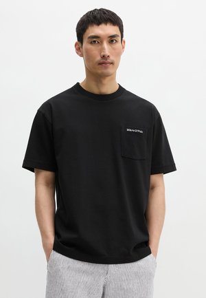 Man draagt zwart T-shirt met korte mouwen en borstzakje met witte tekst, gecombineerd met lichtgekleurde gestreepte broek, staand tegen een witte achtergrond.