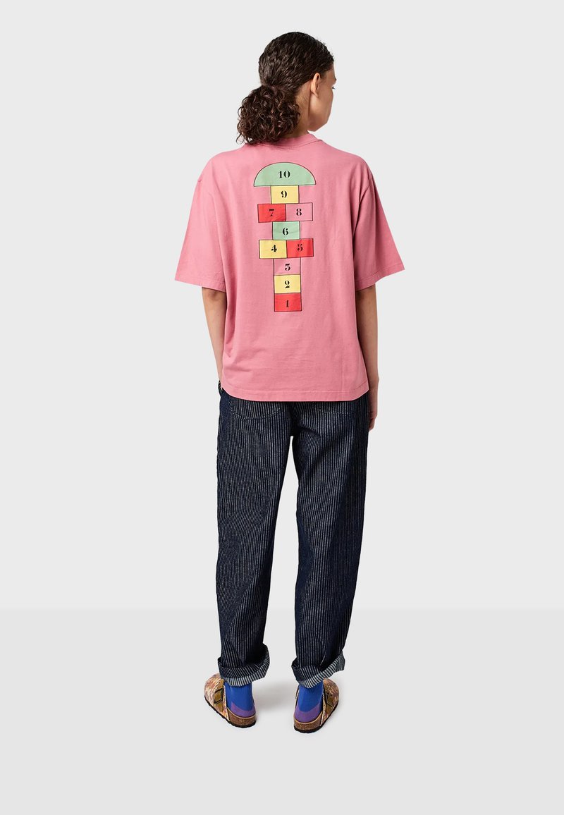 Osoba s kučeravými vlasmi v ružovom tričku s farebným dizajnom skákania na krabicu na zadnej strane, skombinovanom s tmavými pruhovanými nohavicami a šľapkami.