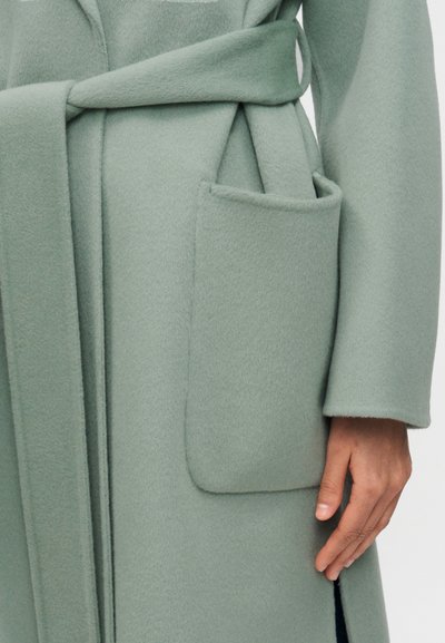 Κοντινή λήψη ενός χεριού που ακουμπά δίπλα στην τσέπη ενός μάλλινου παλτού σε μέντα πράσινο με ζώνη δεμένη γύρω από τη μέση.