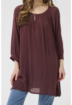 Femme portant une tunique ample à manches longues de couleur mauve foncé avec un petit détail de bouton en forme de trou à l'avant, associée à un jean bleu clair.