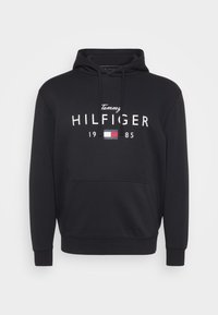 Fekete kapucnis pulóver puha pamut keverékből, elülső zsebpókkal, húzózsinóros kapucnival és merész fehér "Tommy HILFIGER 1985" logóval.