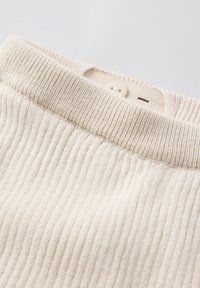 Close-up van een beige geribde trui met een enkele beige knop en een stoffen label aan de binnenkant.