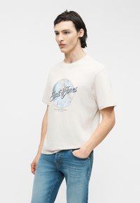 Young man wearing light beige graphic t-shirt with "Jack & Jones" text and blue jeans, standing with one hand in pocket.