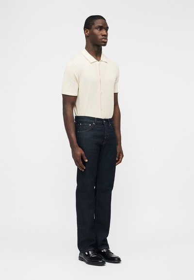 Άνδρας που στέκεται με το πρόσωπο προς τα αριστερά, φορώντας ένα μπεζ κοντομάνικο πλεκτό πόλο, σκούρα μπλε τζιν και μαύρα δερμάτινα παπούτσια, μπροστά σε απλό λευκό φόντο.