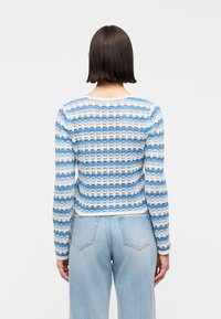 Γυναίκα με κοντά καστανά μαλλιά που φοράει πουλόβερ πλεκτό με ρίγες σε μπλε, λευκό και μπεζ χρώμα και ανοιχτόχρωμο τζιν ψηλόμεσο, κοιτάζοντας προς τα πίσω.