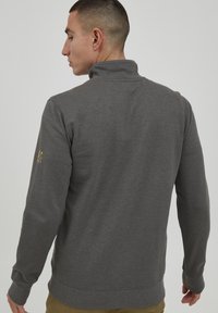 Sudadera gris con un cuello alto, puños y dobladillo acanalados. Presenta un pequeño logo dorado en la manga izquierda. Hecha de un tejido suave y texturizado.