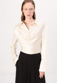 Blusa in satin crema con scollo a cowl, maniche lunghe e chiusura con bottoni. Abbinata a pantaloni neri a vita alta con pieghe.