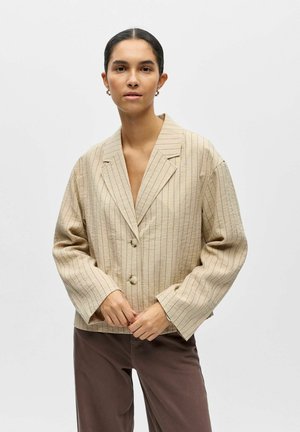 Junge Frau mit dunklen Haaren trägt einen beigen Nadelstreifen-Blazer mit Knöpfen und braune Hosen, steht vor einem schlichten weißen Hintergrund.