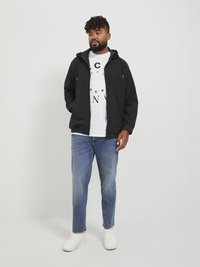 Czarna wodoodporna kurtka z kapturem, pod nią biały t-shirt z grafiką, niebieskie dżinsy i białe sneakersy. Model stoi na neutralnym tle.