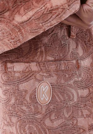 Close-up of textured pink fabric with intricate swirling patterns and a stitched patch featuring a white outlined letter "K".