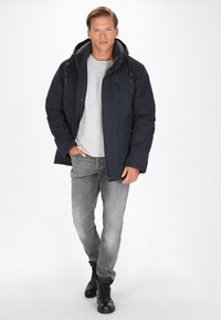 Mørkeblå polstret jakke med grå foring, lynlås foran og justerbar hætte. Bæres over en lysegrå skjorte og parret med grå jeans og sorte støvler.
