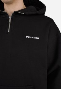 Sweat à capuche noir zippé avec une fermeture éclair en métal et un logo brodé blanc "PEGADOR". Le tissu est texturé, avec une coupe décontractée et une capuche.