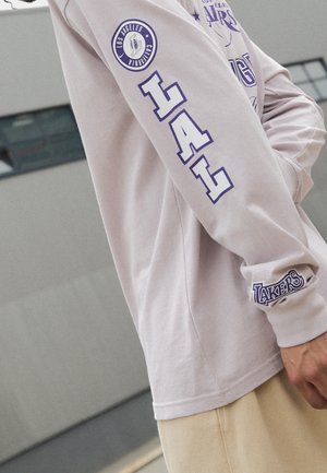 Chemise à manches longues violet clair avec le texte "LA" sur le bras, un patch "Los Angeles California" près de l'épaule, et le texte "Lakers" sur le poignet.