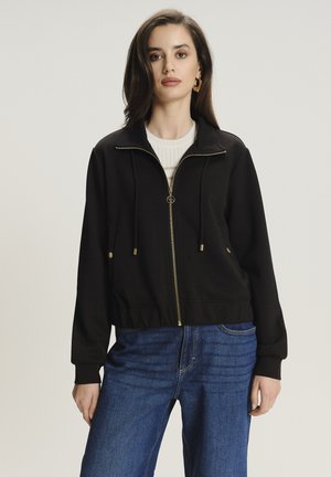 Giacca nera con zip e colletto alto, hardware dorato e coulisse. Abbinata a jeans blu a vita alta. Tessuto: misto cotone.