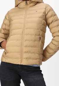 Beżowa puchowa kurtka z kapturem, pikowana faktura, z przodu zamek błyskawiczny i boczne kieszenie. Posiada małe logo na piersi oraz ściągacze na mankietach.